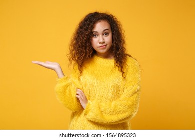 Perplexed confused young african american girl in fur sweater posing isolated on yellow orange background in studio. People sincere emotions lifestyle concept. Mock up copy space. Pointing hand aside