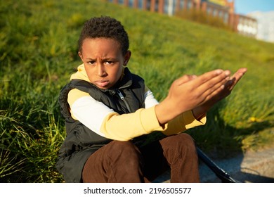 Perplexed And Disappointed Black Kid Boy Sitting Next To His Scooter At City Street Reaching Hands Asking For Help Lost In Unknown Neighborhood, Looking At Camera With Worried And Upset Face