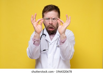 Perplexed Doctor With Poor Eyesight Has Taken Off Glasses And Is Examining Them With A Stupid Expression On Face. Yellow Background.