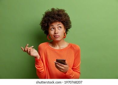 Perplexed hesitant curly haired African American woman shrugs shoulder being not sure uses smartphone device concentrated upwards wears casual orange jumper isolated over green studio background. - Powered by Shutterstock