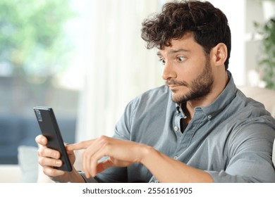 Perplexed man checking cell phone surprising content sitting at home