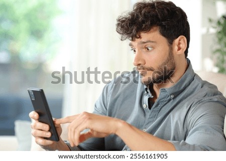 Perplexed man checking cell phone surprising content sitting at home