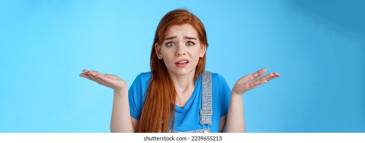 Perplexed uneasy stupified worried redhead woman shrugging, have no idea, frowning nervously, cannot understand what happened, stand clueless unsure, pose blue background. Copy space - Shutterstock ID 2239655213