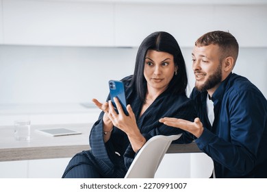Perplexed young couple sitting at kitchen looking at phones screen with puzzled face expression upset by news. Young adult hispanic woman shows to husband, disappointed by economic crisis. Frustration