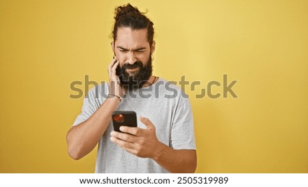 A perplexed young man with a beard looks at his phone against a yellow background.