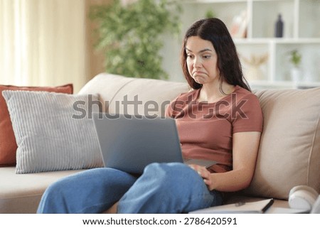 Perplexed young student checking laptop content sitting on a couch at home