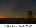 Perseid meteor shower seen outside the city, amidst fields and a hunting stand. Meteor streaks emanate from the radiant point while the sky displays a greenish hue from airglow.