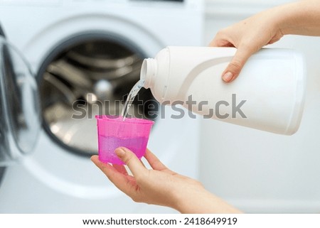 Person adding liquid laundry detergent to the washer, close up. Female hand holding laundry detergent in front of open washing machine