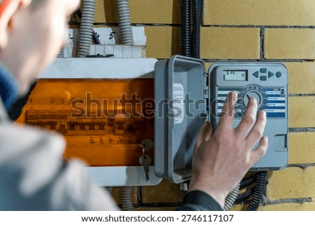 Person adjusting irrigation controller for automatic watering system, managing sprinkler zones via digital control panel mounted on brick wall.