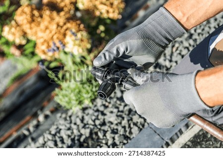 A person adjusts an irrigation tool while standing on a garden path next to blooming flowers in bright daylight.
