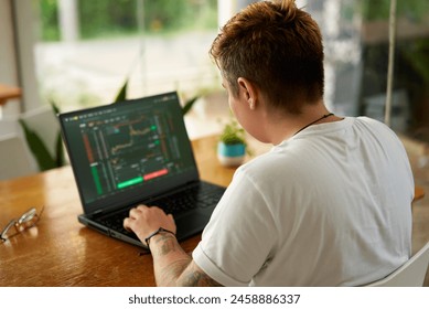 Person analyzes financial markets on laptop with charts. Gender inclusive investor focused, trades in modern home office. Casual workday fintech professional. Analysis, strategy comfy surroundings. - Powered by Shutterstock