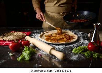 A person in an apron spreads tomato sauce on fresh pizza dough. Surrounding ingredients include tomatoes and herbs, creating a cozy, home-cooked atmosphere. - Powered by Shutterstock