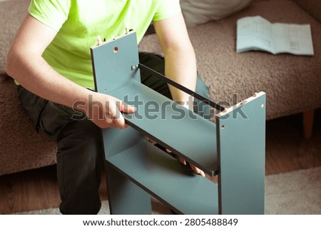 Person assembling a gray bookshelf while seated on a couch in a living room, with an instruction manual visible on the surface nearby