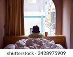 A person in bed looking out of the hotel room window admiring cityview. Selective focus, solo travelling.