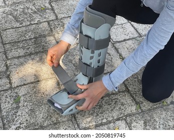 Person bending down in the street adjusting the velcro straps of the orthopedic walking boot, ideal for people with leg injuries such as tibia or fibula fracture. Patient walking down the street. - Shutterstock ID 2050819745