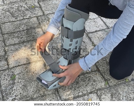 Person bending down in the street adjusting the velcro straps of the orthopedic walking boot, ideal for people with leg injuries such as tibia or fibula fracture. Patient walking down the street.