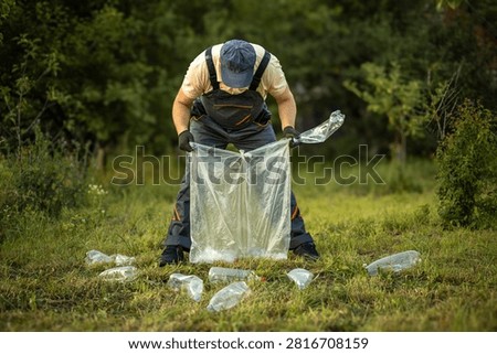A person bends down to pick up plastic bottles in a grassy area. The sun is shining as they clear the litter from the environment. This activity promotes cleanliness and care for nature.