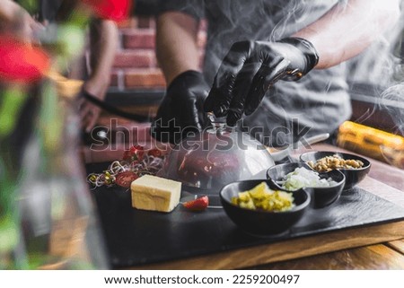 Person in black gloves smoking beef tartare under transparent lid served on black wooden board with garnishes on the side. Polish cuisine. Horizontal indoor shot. High quality photo