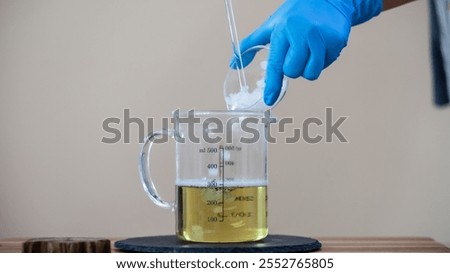 A person in blue gloves adds a solid substance into a measuring cup containing a yellow liquid, indicating a chemical or scientific experiment.