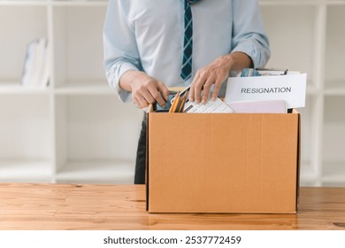 A person in a blue shirt and tie is unpacking documents from a cardboard box on a wooden desk in a well-lit office space. - Powered by Shutterstock