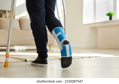 Person With Broken Foot Walking On Crutches. Young Man Wearing Ankle Support Brace Or Fracture Fixator With Adjustable Straps Walking In Living Room At Home. Close Up Of Human Feet. Leg Injury Concept