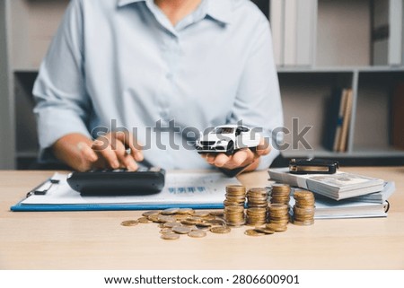 Person calculating auto financing while holding a white toy car, with calculator, cash, and coins on desk, representing car loan budgeting, vehicle purchase cost, and finance management.