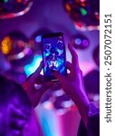 A person captures a vibrant nightclub scene with their smartphone, focusing on reflective disco balls under colorful neon lights. The image emphasizes the lively and immersive atmosphere of the event.