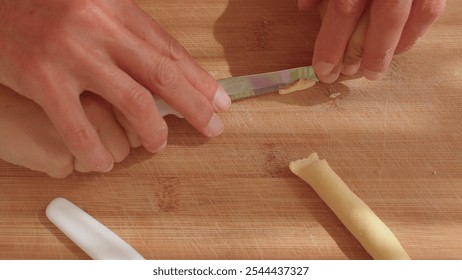 A person is carefully cutting piece of dought with a knife on a sturdy wooden cutting board. In the Puglia region, a traditional handmade pasta with a round shape is called orecchiette. - Powered by Shutterstock