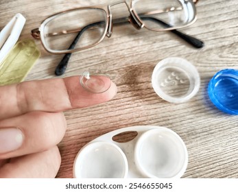 A person carefully holds a contact lens on their fingertip next to glasses and lens care products on a table. - Powered by Shutterstock