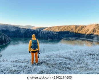 A person carrying a heavy backpack is standing on a snowy hilltop, taking in the breathtaking views of a serene lake below while appreciating the stunning beauty of nature surrounding them completely - Powered by Shutterstock