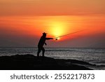 A person casting a fishing line against a vibrant sunset over the ocean.