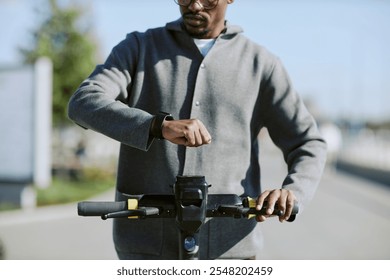 Person in casual attire checking smartwatch while standing on electric scooter, outdoors with blurred background suggesting movement and activity in urban area - Powered by Shutterstock