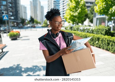 A person checks a package while standing on the sidewalk. The setting is a busy city street with tall buildings and greenery. It is daytime and people can be seen in the background.