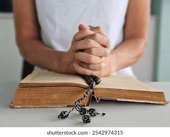 A person with clasped hands praying over an open book with prayer beads on a table in a serene setting - Powered by Shutterstock