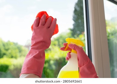 A person is cleaning a window using a red cloth and a spray bottle. The sun is shining, and there is fresh greenery visible outside.