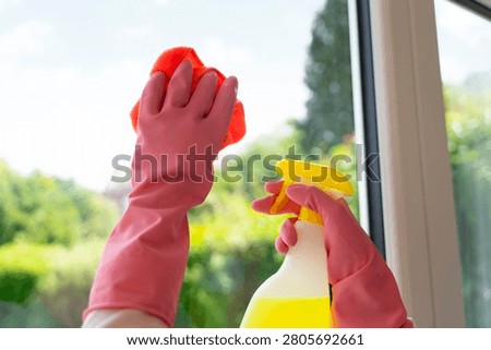 A person is cleaning a window using a red cloth and a spray bottle. The sun is shining, and there is fresh greenery visible outside.