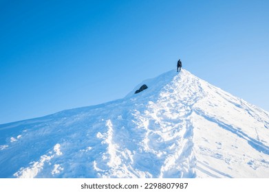 Person clibming up snowy mountain peak. Blue sky in afternoon. Hiker or climber snowshoeing up to top of big rock.