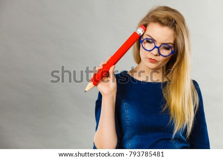 Person confused thinking seeks a solution. Pensive thoughtful student girl or business woman female teacher wearing nerdy glasses coming up with an idea, holding big red pencil. Studio shot on grey.