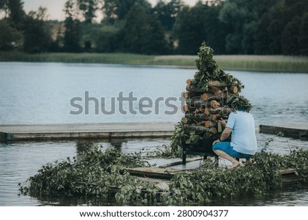 Person constructs a natural decoration using logs and foliage on a floating platform in a calm lake, with trees and greenery in the background