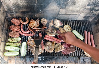 Person Cooking Assorted Meat And Vegetables On An Outdoor BBQ Using Metal Utensils In A High Angle First Person POV Looking Don At The Grill