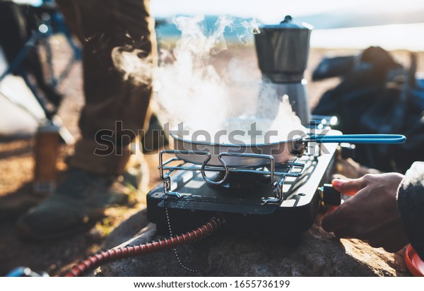 person cooking food and hot tea in\
nature camping outdoor, cooker prepare breakfast picnic on metal\
gas stove, tourism recreation outside; campsite\
lifestyle