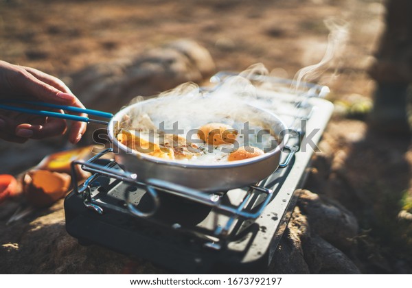 person cooking\
fried eggs in nature camping outdoor, cooker prepare scrambled\
omelette breakfast picnic on metal stove, tourist on recreation\
outside; campsite\
lifestyle