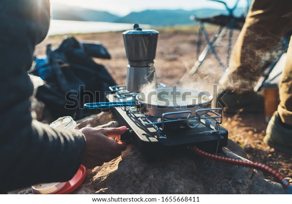 person cooking hot drinks in nature\
camping outdoor, cooker prepare breakfast picnic on metal gas\
stove, tourism recreation outside; campsite\
lifestyle