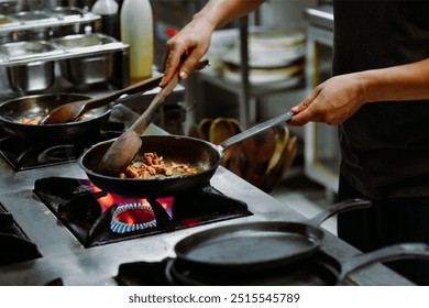 a person cooking in a professional kitchen, stirring ingredients in a frying pan over a gas flame. The cook uses a wooden spoon and metal spatula to sauté what appears to be pieces of meat or vegetabl - Powered by Shutterstock