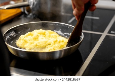 person is cooking scrambled eggs in a pan on a stovetop, stirring with a wooden spatula. The steam rising from the pan adds to the enticing moment, evoking the warmth and aroma of freshly cooked food - Powered by Shutterstock