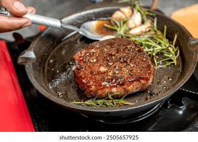 A person is cooking a tenderloin steak in a pan with herbs and spices. The steak is brown and he is well-cooked