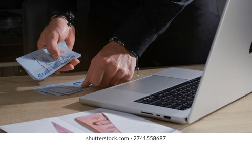 Person counts cash while using a laptop on a wooden table in an indoor setting during daytime