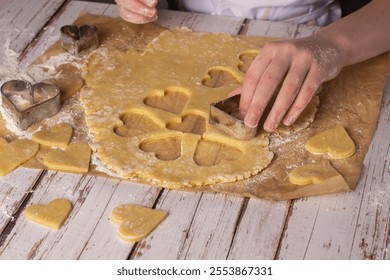 A person cuts out heart shapes from cookie dough using metal cookie cutters. Flour covers the wooden table, creating a messy yet enjoyable baking experience in a cozy kitchen setting. - Powered by Shutterstock