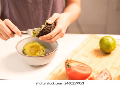 A person is cutting an avocado and placing it in a bowl. There are also tomatoes and limes on the table - Powered by Shutterstock