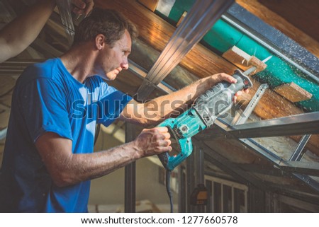 A person is cutting a board of wood next to a window with a reciprocating saw. Sawdust is flying around. Detail of a man cutting wooden board with strong back light to emphasize saw dust.
